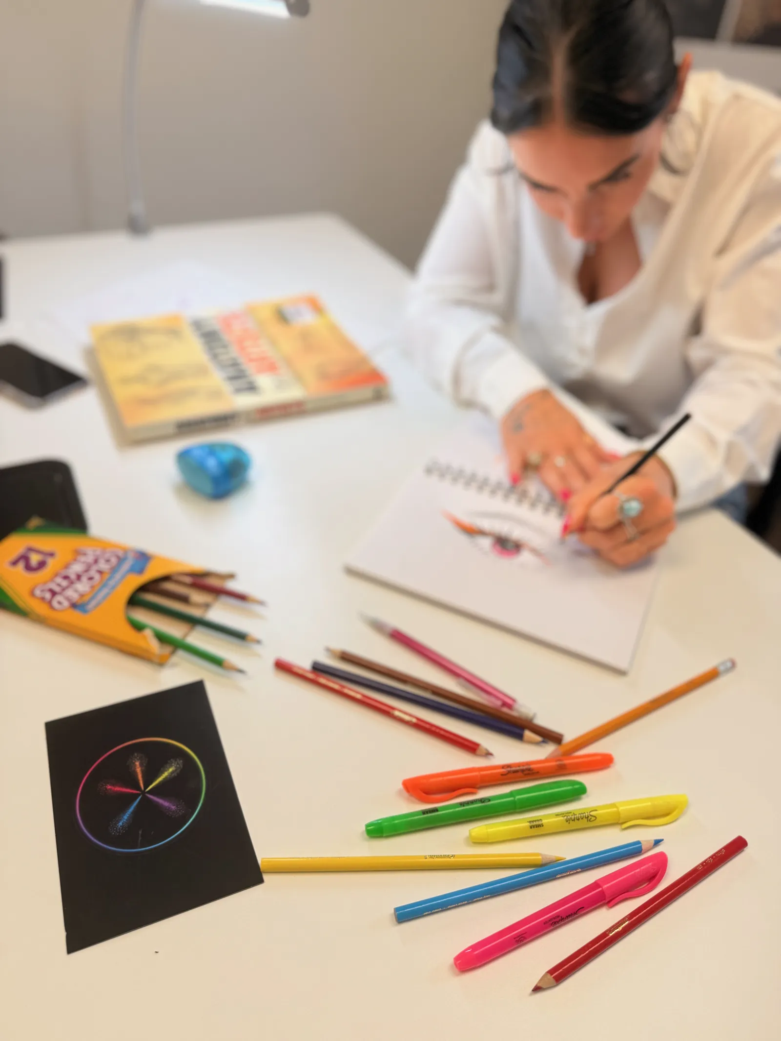 Taylor drawing an eye in her sketchbook, colored pencils and markers on the table